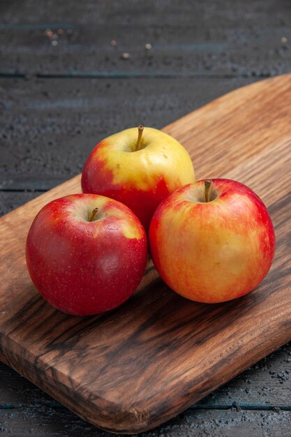 Vista lateral de perto frutas na tábua, uma maçã vermelha e duas maçãs amarelo-vermelhas em uma tábua marrom na mesa cinza