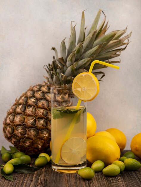 Vista lateral de suco de limão rico em vitaminas em um copo com limões de abacaxi e kinkans isolados em uma mesa de madeira sobre uma superfície branca