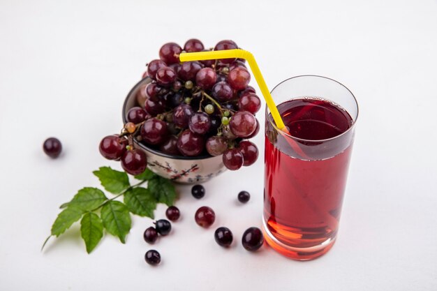 Vista lateral de suco de uva preta com tubo de bebida em vidro e tigela de uvas vermelhas com folhas em fundo branco