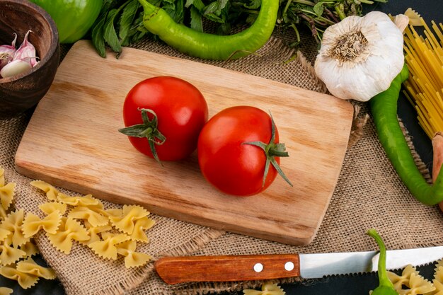 Vista lateral de tomates em uma tábua de cortar com faca de alho e pimenta e hortelã em um guardanapo bege