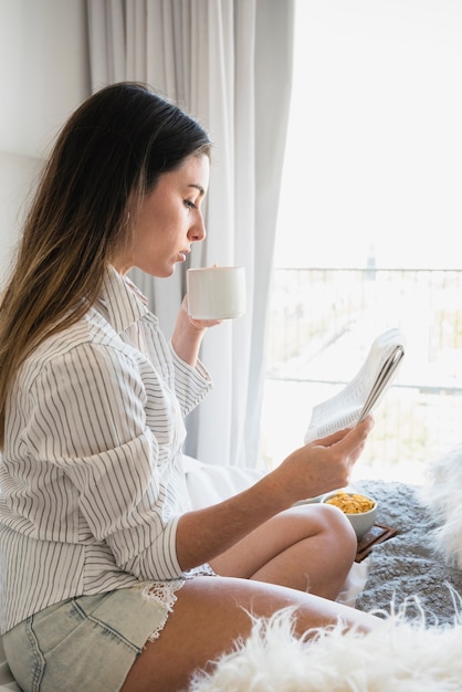 Vista lateral, de, um, assento mulher, cama, bebendo, a, café, enquanto, jornal leitura