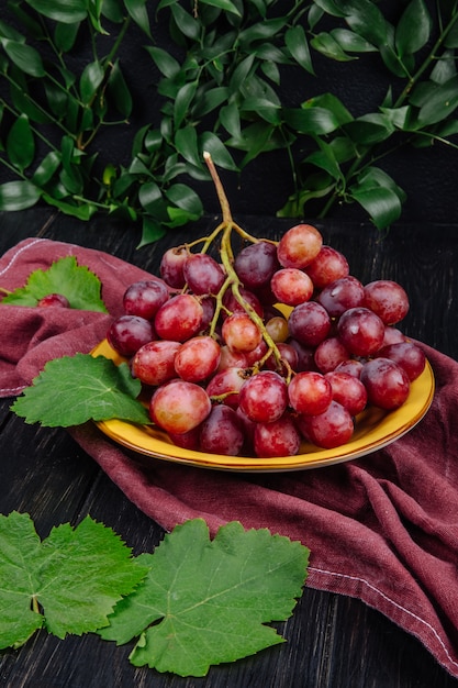 Foto grátis vista lateral de um cacho de uvas doces frescas em um prato e folhas de uva verde na mesa de madeira