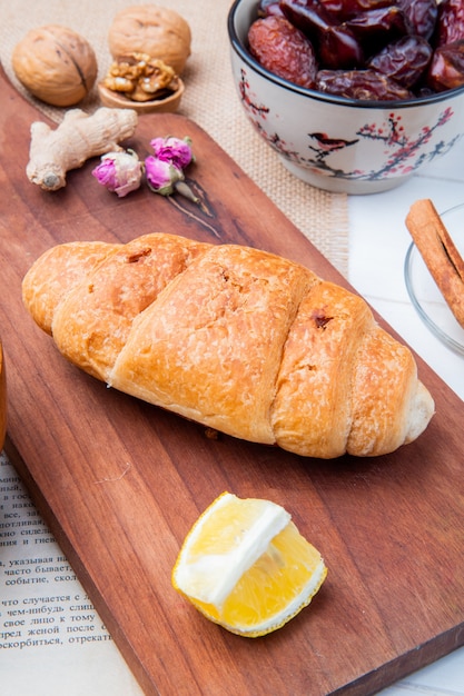 Vista lateral de um croissant com um pedaço de limão em uma tábua de madeira e com tâmaras secas doces em uma tigela e nozes em madeira branca