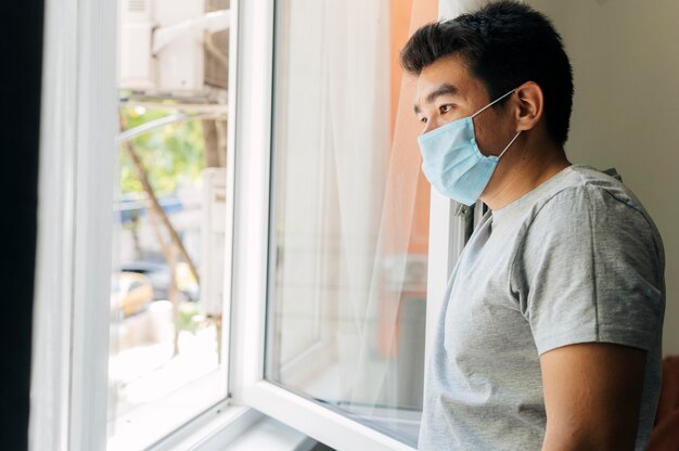 Vista lateral de um homem com máscara médica em casa durante a pandemia, olhando pela janela