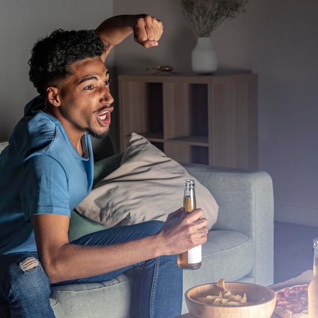 Vista lateral de um homem torcendo para a tv em casa com cerveja e petiscos