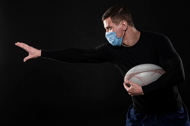 Vista lateral de um jogador de rugby com máscara médica e bola