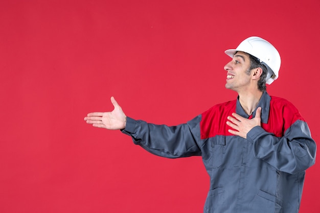 Vista lateral de um jovem trabalhador confiante de uniforme com capacete apontando algo no lado direito na parede vermelha isolada