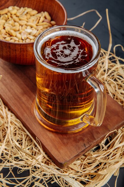 Vista lateral de uma caneca de cerveja e amendoins em uma tigela na placa de madeira com palha no rústico