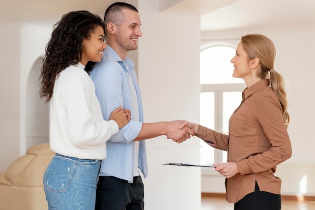 Vista lateral de uma corretora de imóveis apertando as mãos de um casal para um novo contrato