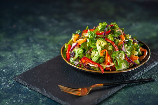 Vista lateral de uma deliciosa salada vegan com ingredientes frescos em um prato e um garfo em uma tábua preta sobre fundo azul