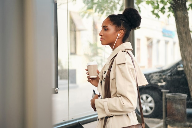 Vista lateral de uma garota afro-americana casual em um elegante casaco de trincheira com café para ir e celular assistindo a rota no ponto de ônibus