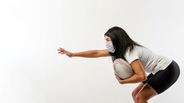 Vista lateral de uma jogadora de rugby com bola e máscara médica