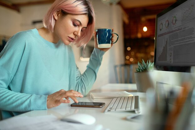 Vista lateral de uma jovem blogueira elegante de cabelo rosa sentado à mesa em frente ao computador desktop