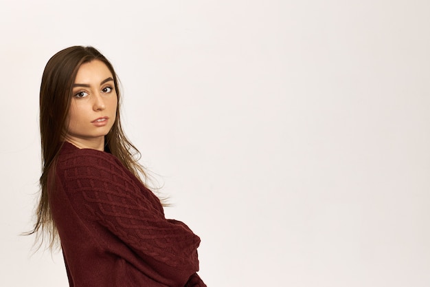 Vista lateral de uma jovem mulher de cabelos escuros, elegante e confiante em um suéter aconchegante e aconchegante, cruzando os braços sobre o peito e sorrindo para a câmera, posando contra um fundo branco com espaço de cópia