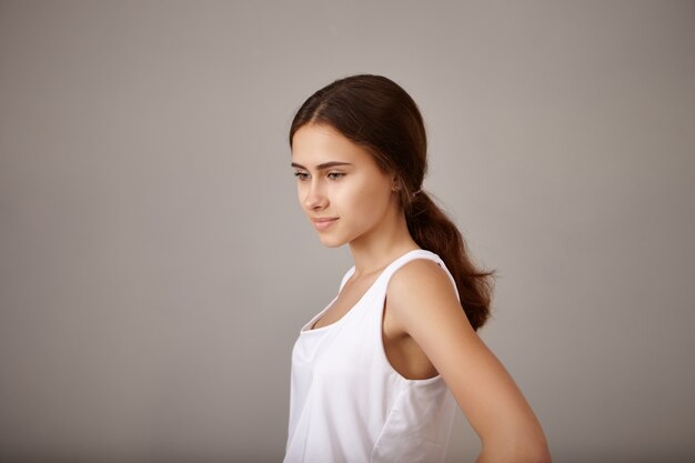 Vista lateral de uma jovem pensativa, pensativa, de vinte anos, vestida de top branco, posando isolada na parede em branco do copyspace, olhando para baixo, sonhando acordada, tendo profunda expressão de pensamentos