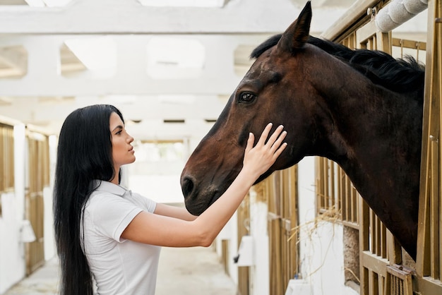 Vista lateral de uma linda mulher com grande cavalo marrom no estábulo