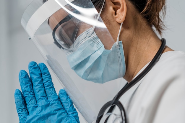 Vista lateral de uma médica com protetor facial e máscara médica orando
