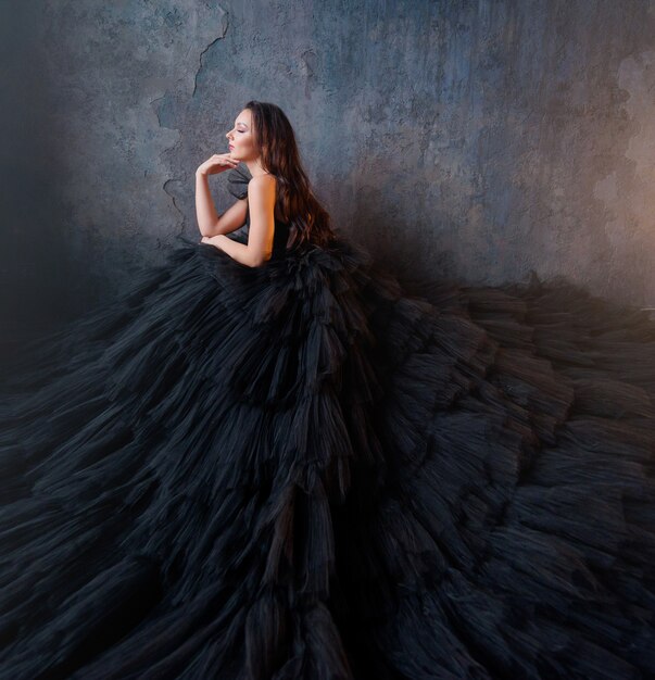Vista lateral de uma morena com um vestido preto exuberante, sentada em um fundo da parede