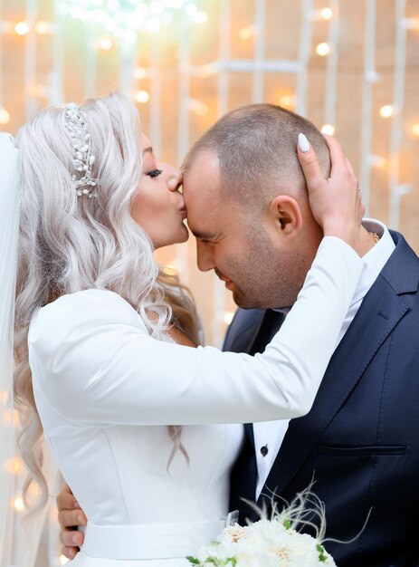 Vista lateral de uma mulher adorável que tem um lindo penteado com joias vestidas de vestido branco, abraçando o marido e beijando
