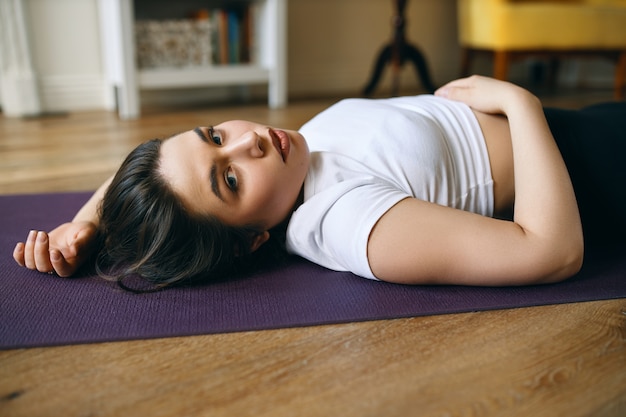 Vista lateral de uma mulher caucasiana atraente jovem plus size em roupas esportivas, deitada de costas no tapete de ioga após o treino, olhando para a câmera com expressão facial cansada.