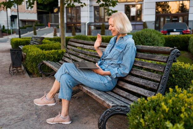 Vista lateral de uma mulher mais velha ao ar livre no banco acenando para um laptop