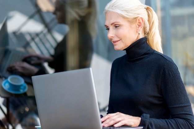 Vista lateral de uma mulher mais velha trabalhando em um laptop ao ar livre enquanto saboreia um café