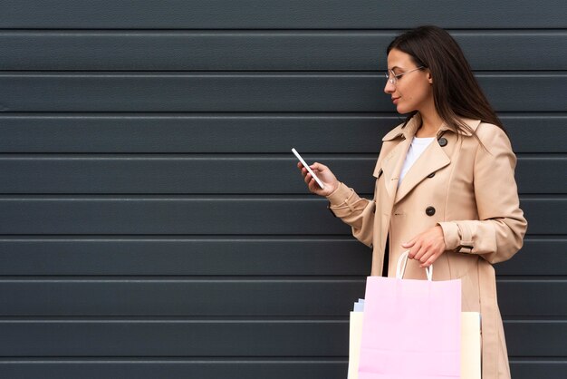 Vista lateral de uma mulher olhando para o smartphone enquanto segura sacolas de compras
