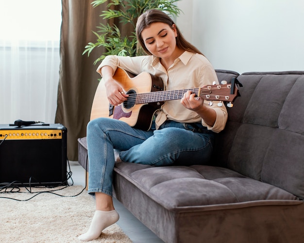 Vista lateral de uma musicista em casa tocando violão enquanto está sentada no sofá