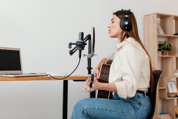 Vista lateral de uma musicista gravando música com microfone enquanto toca violão em casa