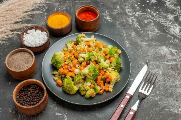 Vista lateral de uma refeição saudável com brocoli e cenoura em uma placa preta e especiarias na mesa cinza