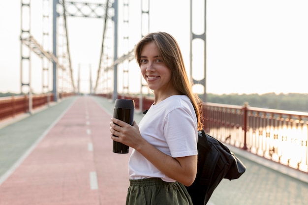 Vista lateral de uma sorridente mulher viajando segurando uma garrafa térmica na ponte