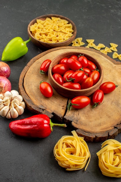 Vista lateral de uma tigela de longe na tigela de tomate na tábua de cortar macarrão e cebola, pimentão e alho na mesa