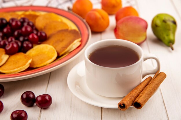 Vista lateral de uma xícara de chá e canela no pires com panquecas e cerejas no prato e damascos de pêssego pêra no fundo de madeira