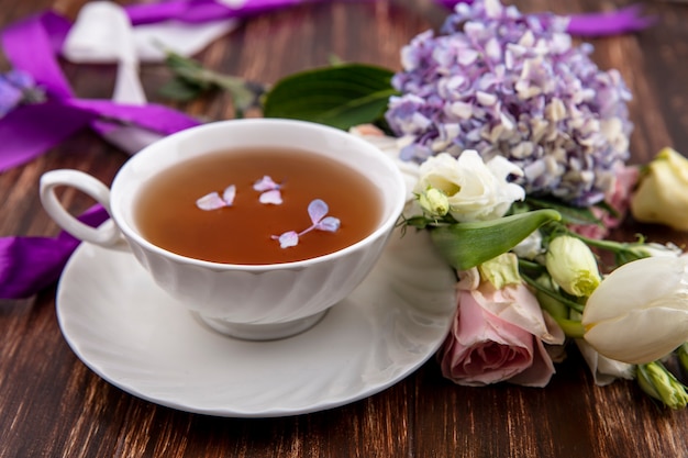 Vista lateral de uma xícara de chá no pires e flores com fitas no fundo de madeira