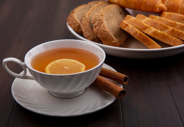 Vista lateral de uma xícara de toddy quente com limão dentro e canela no pires com pães fatiados em um prato com fundo de madeira