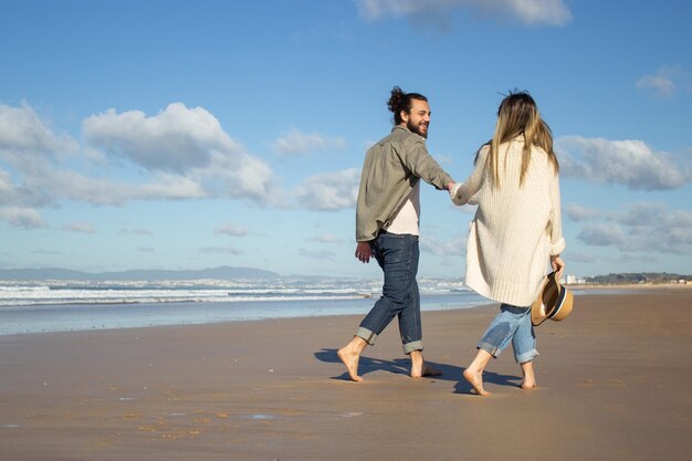 Vista lateral do casal caucasiano nas férias de verão. Homem barbudo e mulher em roupas casuais andando na praia, de mãos dadas. Amor, viajar, conceito de ternura