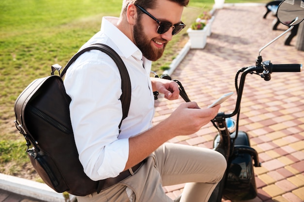 Vista lateral do homem barbudo sorridente em óculos de sol, sentado na moto moderna ao ar livre e usando o smartphone