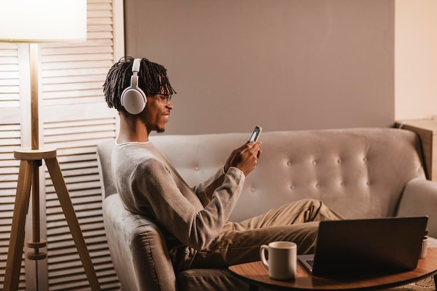 Foto grátis vista lateral do homem em casa no sofá usando smartphone e fones de ouvido