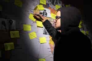 Foto grátis vista lateral do homem planejando ataque de hackers no quarto escuro. homem escrevendo na parede com adesivos, fotografias e fios vermelhos. planejamento, conspiração, conceito de hacking