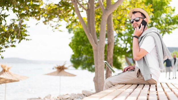 Vista lateral do homem que trabalha na praia com laptop e smartphone