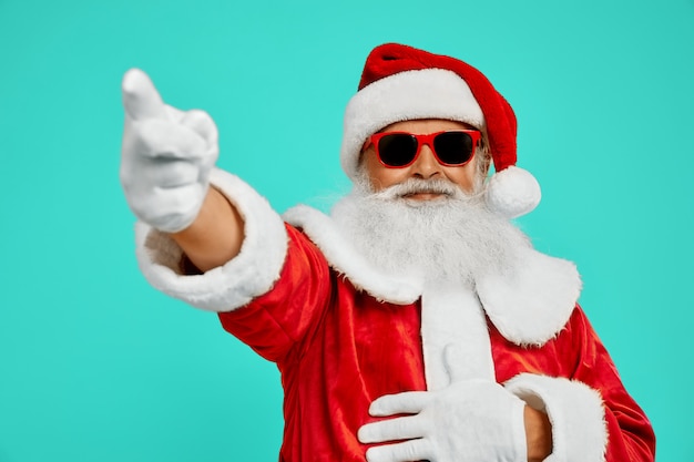 Vista lateral do homem sorridente em traje vermelho de Papai Noel. Retrato isolado de homem mais velho com barba branca longa em óculos de sol, apontando para fora