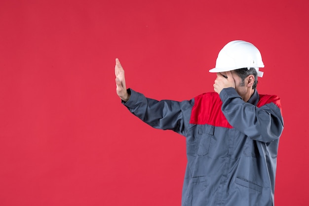Foto grátis vista lateral do jovem arquiteto de uniforme com capacete e fechando os olhos na parede vermelha isolada