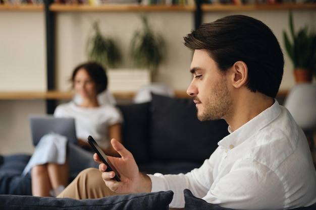 Vista lateral do jovem empresário bonito sentado no sofá com o colega no fundo enquanto sonhadoramente usando o celular no espaço de coworking moderno