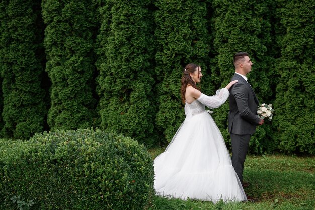 Vista lateral elegante noiva jovem em vestido de noiva inchado em pé atrás do noivo bonito que segurando o buquê de flores posando juntos no jardim Noivas casal e momento de ternura