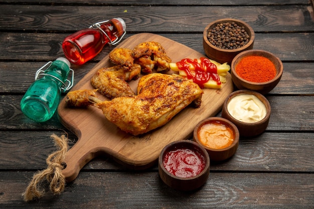 Vista lateral em close-up de frango com coxa e asas de frango com batata frita e ketchup na tábua de cortar ao lado dos molhos de pimenta preta, especiarias e garrafas vermelhas e azuis