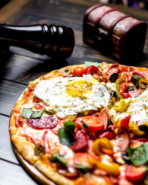 Vista lateral estreita pizza misturada com tomates azeitonas pimentão ovos salsichas no quadro um livro uma faca e um garfo na mesa