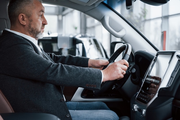 Vista lateral. Homem de negócios sênior em roupas oficiais, tentando novo carro de luxo no salão de automóvel