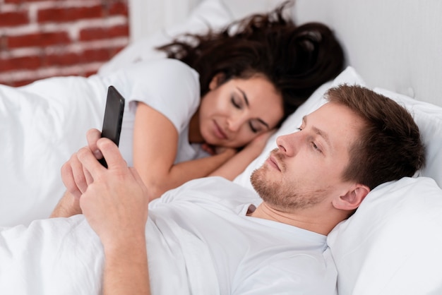 Vista lateral homem verificando seu telefone ao lado de namorada dormindo
