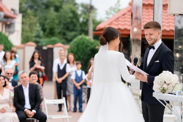 Foto grátis vista lateral sorridente noivo homem com microfone segurando a mão da noiva jura um juramento na cerimônia de casamento ao ar livre convidados felizes no fundo apreciando o lindo casal esposa elegante no vestido inchado