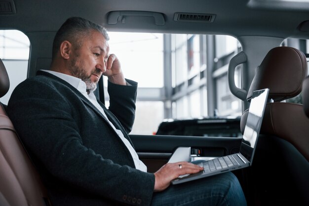 Vista lateral. Trabalhando em uma traseira do carro usando o laptop de cor prata. Homem de negócios sênior